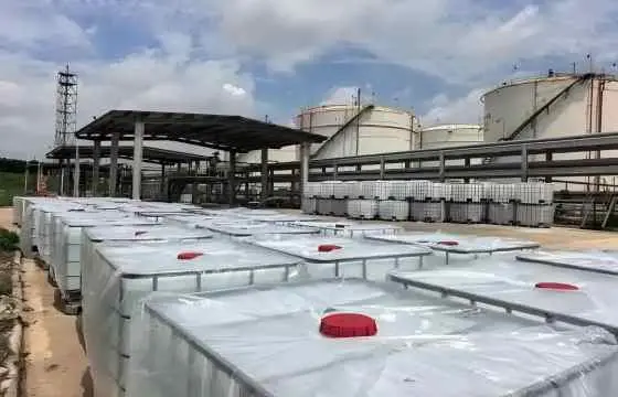 成片包裹着塑料膜、带有红色顶盖的吨桶整齐摆放在化工储罐厂区的露天场地，背景可见大型白色罐与管道构架，甲醇主要应用于甲醛、醋酸、二甲醚、烯烃、汽油添加剂、溶剂、防冻液及燃料等行业。